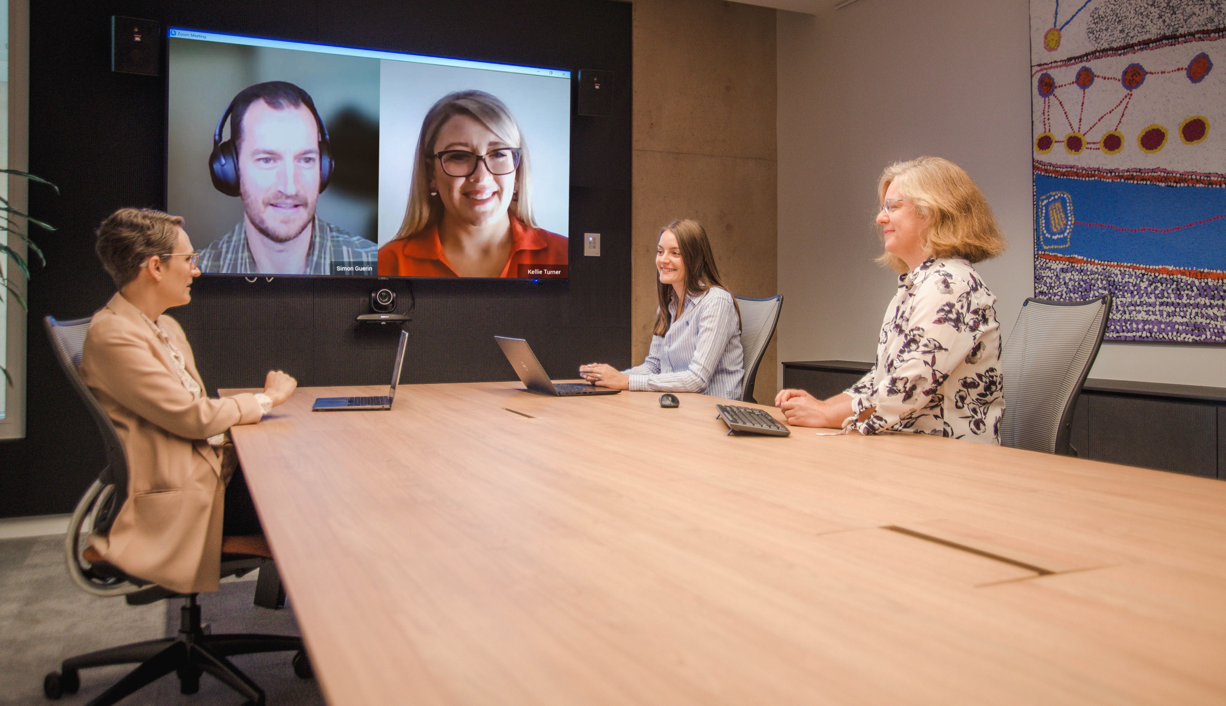Dr Angie Knaggs, Lara Harbers and Dr Sabine Matook sitting at a big conference table talking to Simon Guerin and Kellie Turner via Zoom. Simon and Kellie's Zoom windows are projected on the big screen behind them.