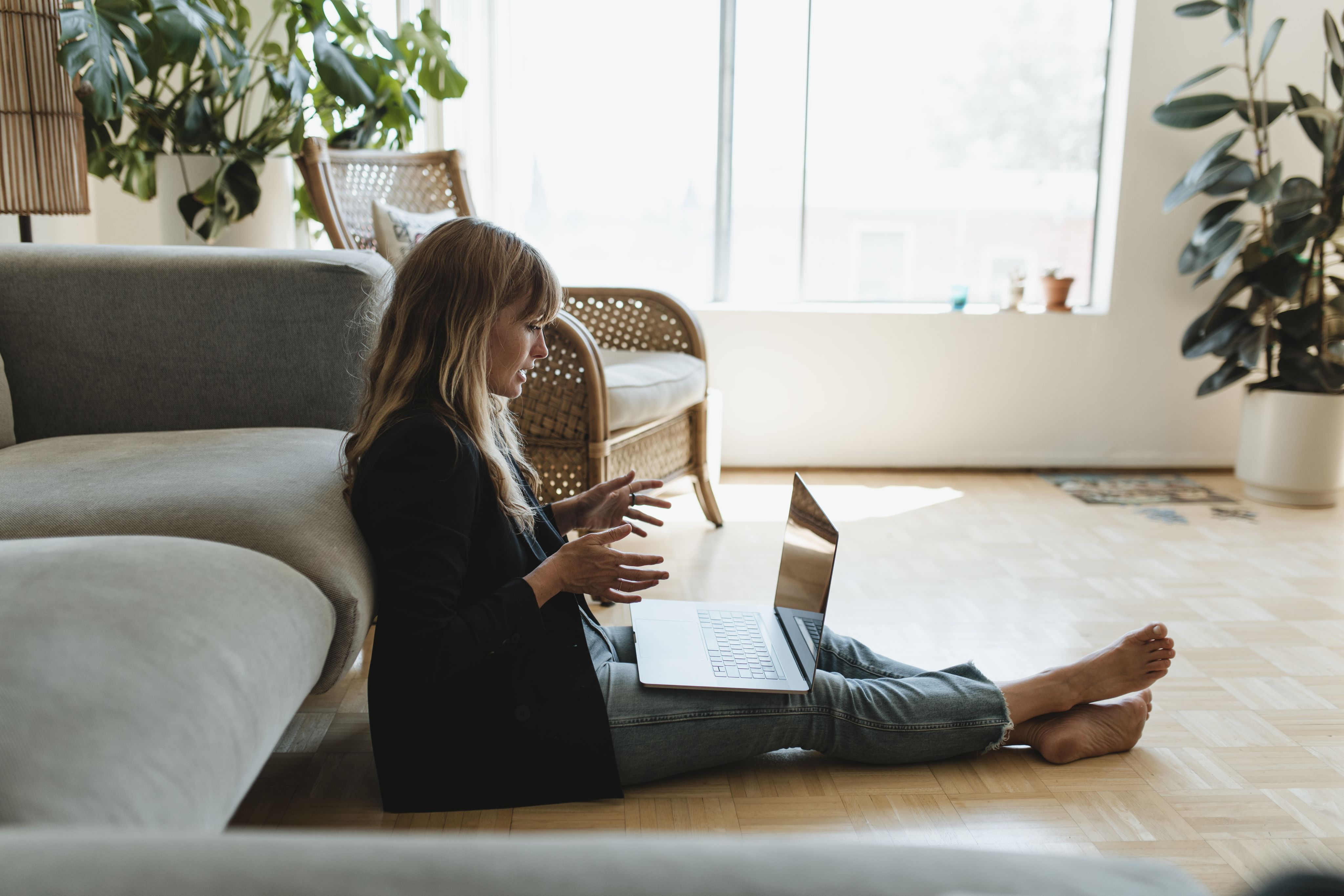 A blonde woman sitting on the floor in front of her couch, gesturing at her laptop