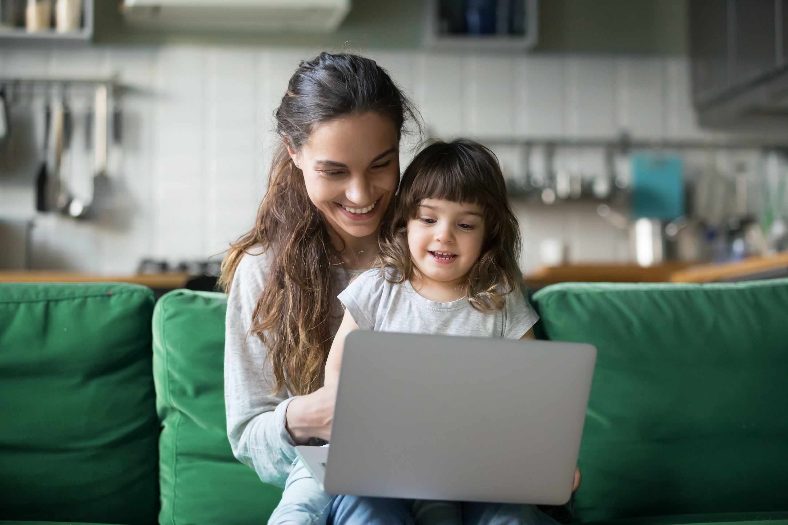 Happy laughing mother and preschool daughter using laptop together on a green couch.