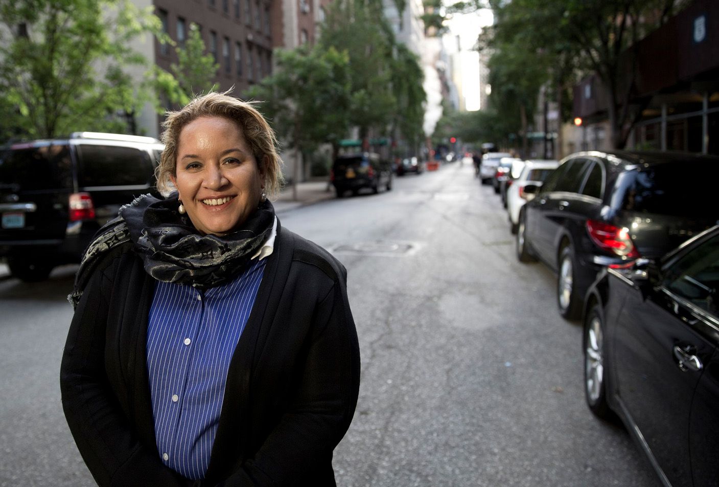 An Aboriginal woman smiles at the camera. She is standing in a street lined with cars and trees. She is wearing a dark coat with a blue shirt, a scarf and pearl earrings.
