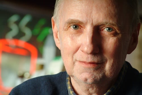 A Caucasian man looking seriously into the camera. He is older, with grey hair. He is wearing a blue jumper.