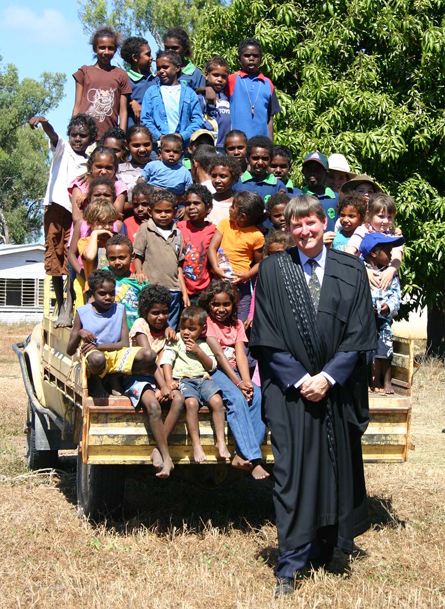 A white man stands in formal robes, smiling, in front of a ute. Behind him are twenty or so Indigenous and local children