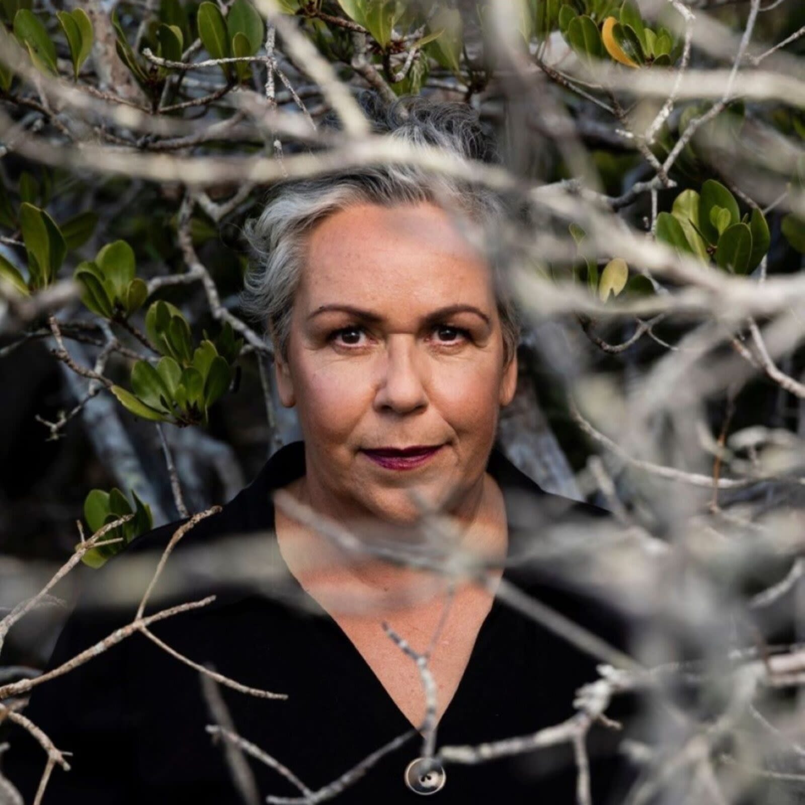 An Indigenous woman looks seriously at the camera. She is standing amidst a tree, and branches obscure parts of the photograph. She has short, grey hair and lipstick and is wearing a black shirt.