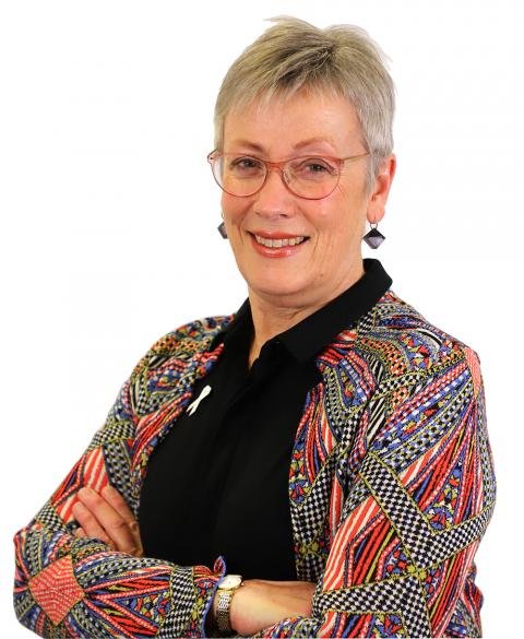 A headshot of a Caucasian woman wearing a colourful cardigan. She has red glasses and short grey hair. Her arms are crossed and she is smiling at the camera.