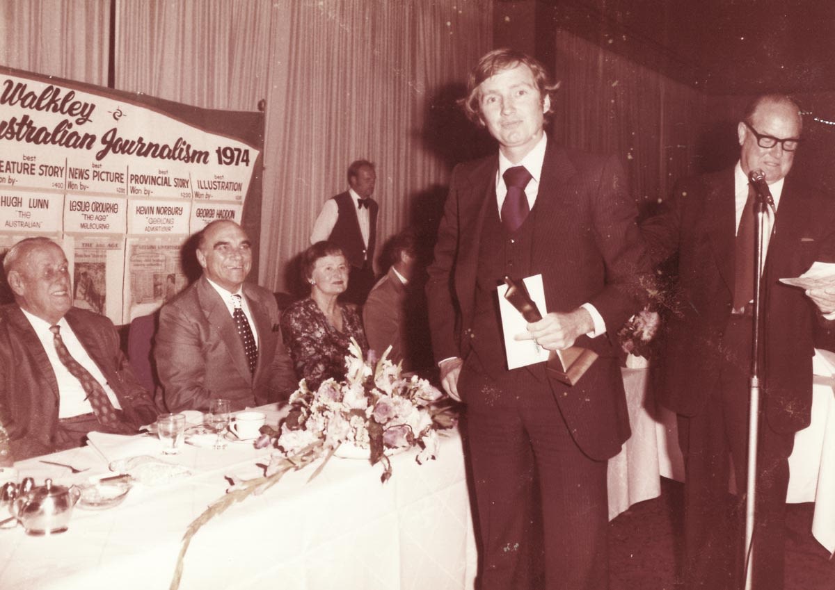 An old sepia photo of a young man holding an award and smiling into the camera. Another man is announcing something next to him, and a row of people sit at a table next to the man holding the award, looking up at him and smiling. A sign behind them reads 'Walkley - Australian Journalism 1974'.