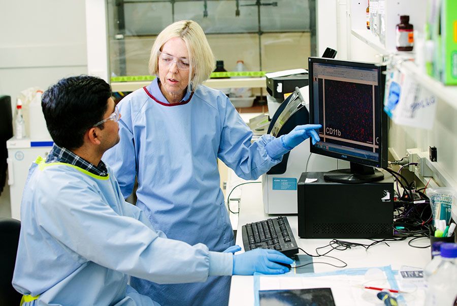 A woman in a lab coat points to a computer screen. She is wearing gloves and protective glasses. She is talking with the man at the computer, who also wears personal protective equipment, and is using the computer. They are in a laboratory.
