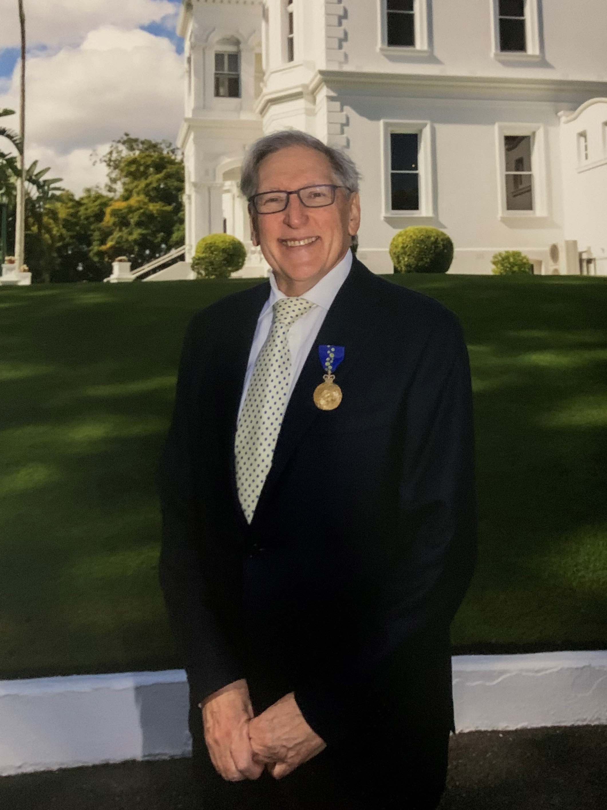 A portrait of an older man wearing a formal suit and a medal. He stands outside a grand white building, in front of a grassy lawn.