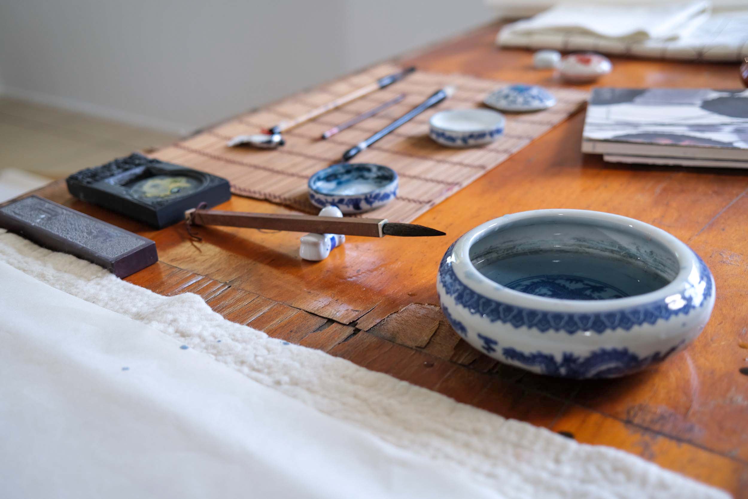 An old wooden table with various art supplies including an ink grinder, ceramic bowls, and a brush resting on a brush holder.