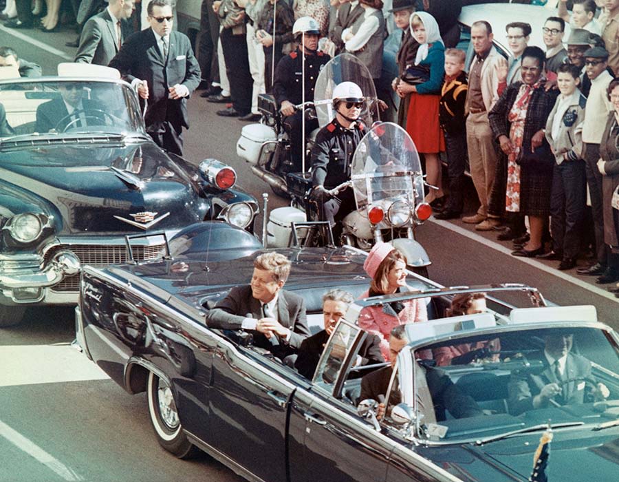 US President John F. Kennedy and his wife, Jackie Kennedy, ride in a topless black car as crowds gather on the sidelines. A police escort on a motorcycle is beside them.