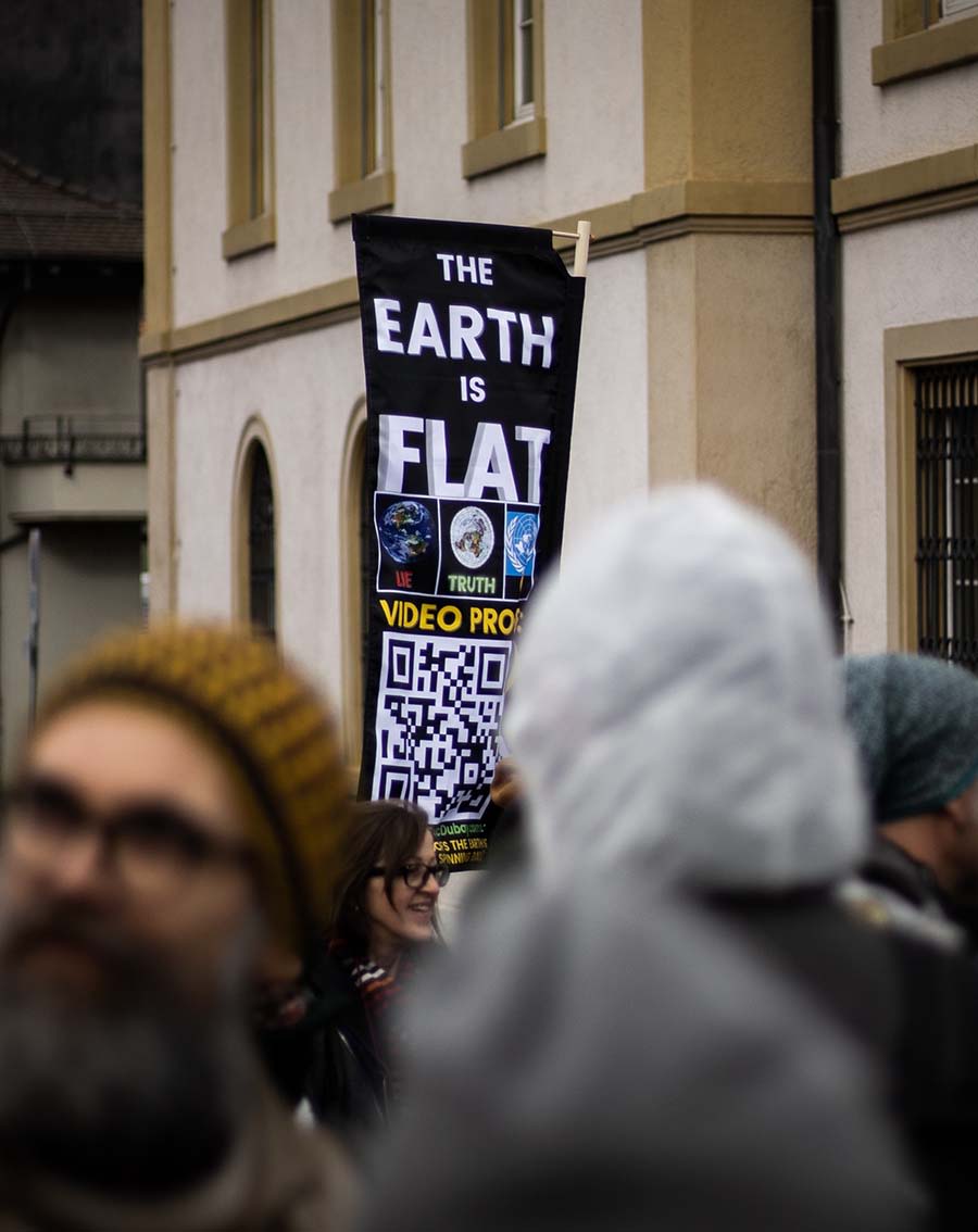 A blurred foreground of heads behind which a clear poster reading 'the earth is flat - video proof' written on it with a big QR code.