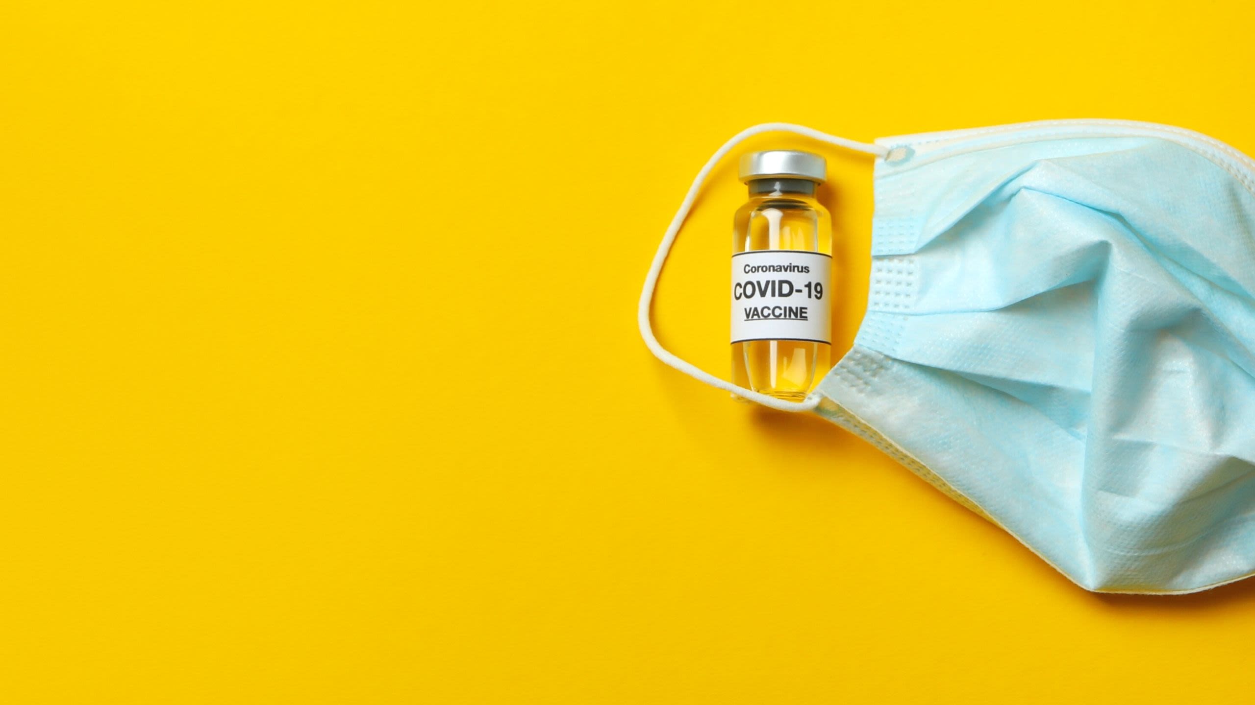 Vial of Covid-19 vaccine and blue mask on yellow background