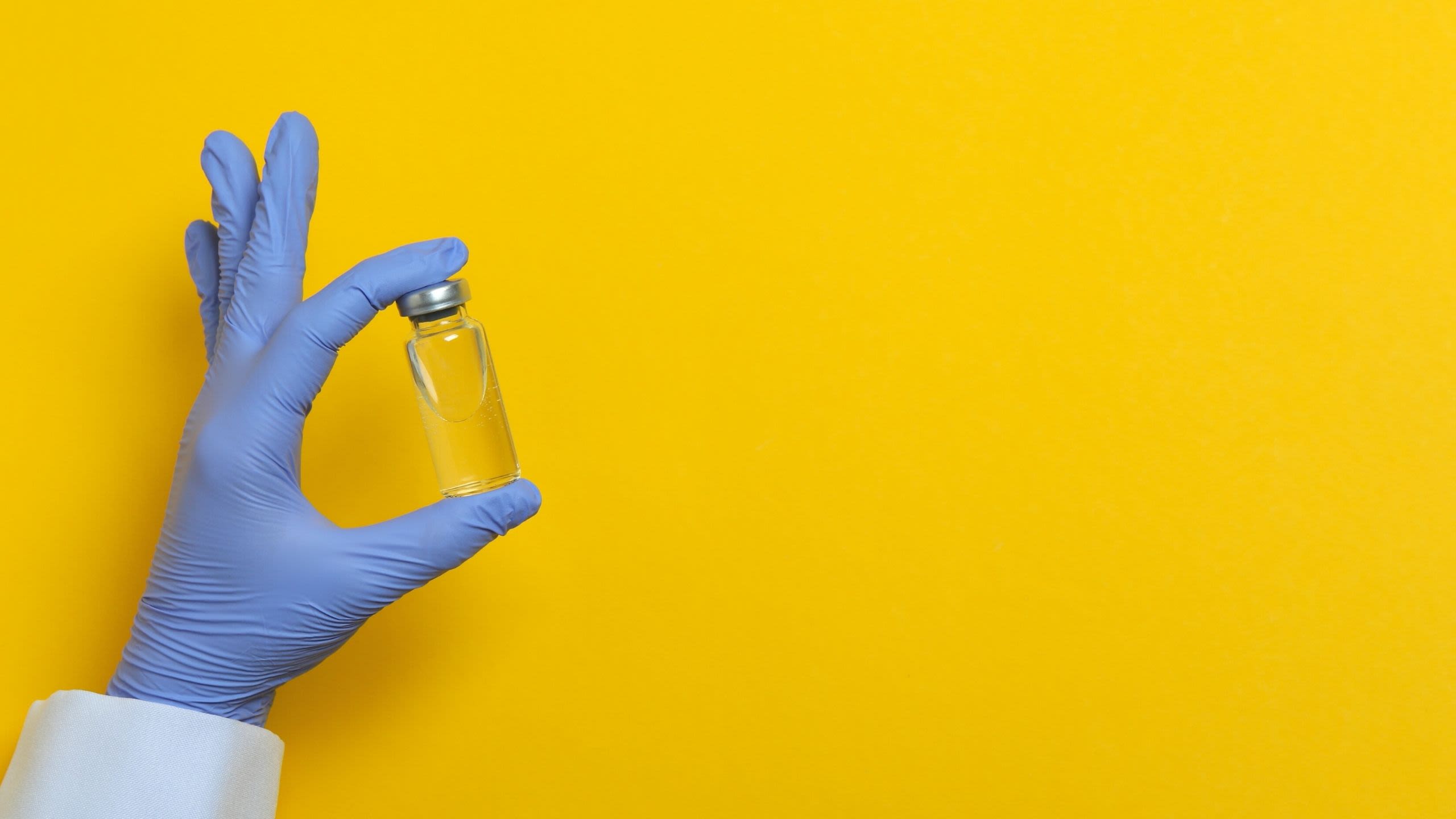 Hand in blue medical glove holding a Covid-19 vaccine against a yellow background