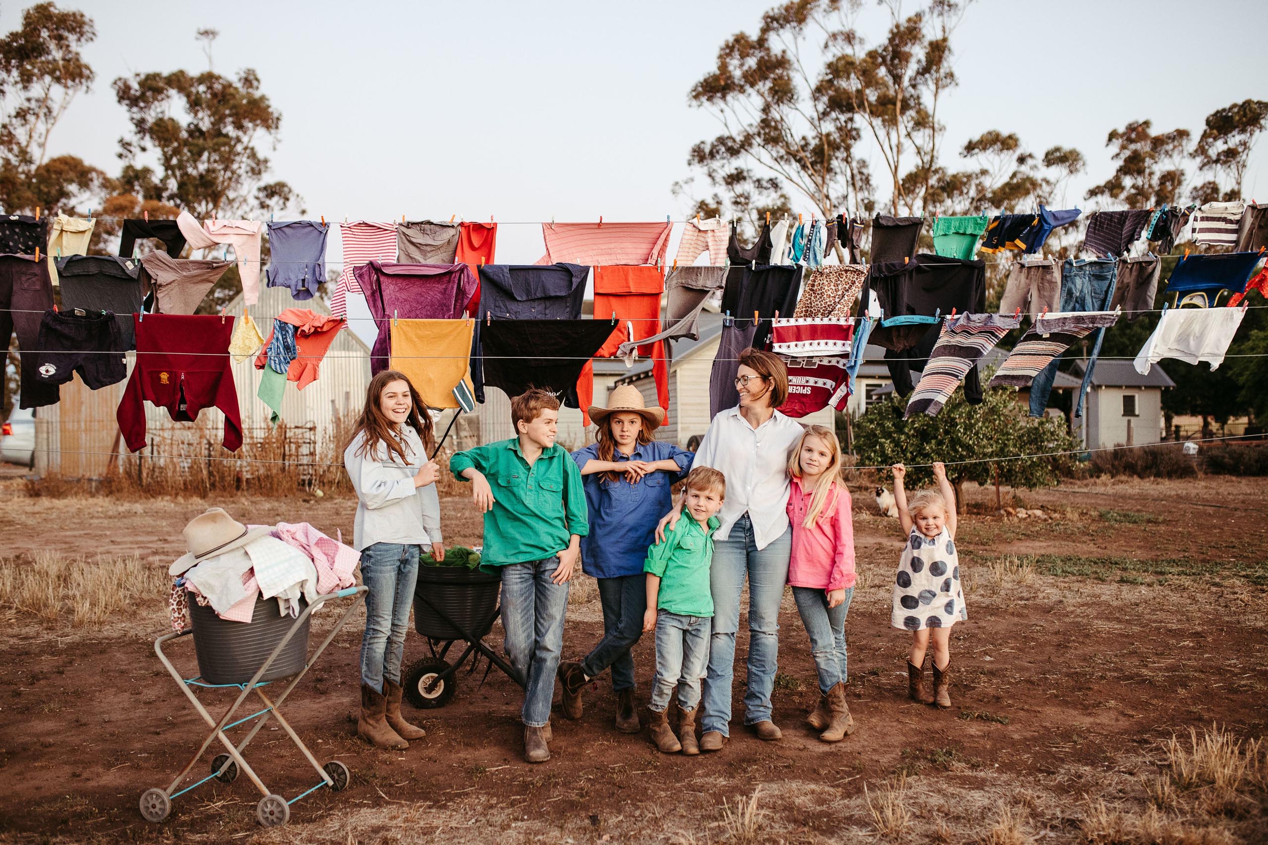 Family in front of a clothes line. There are six children and one mother and three rows of washing hanging on the clothes line.