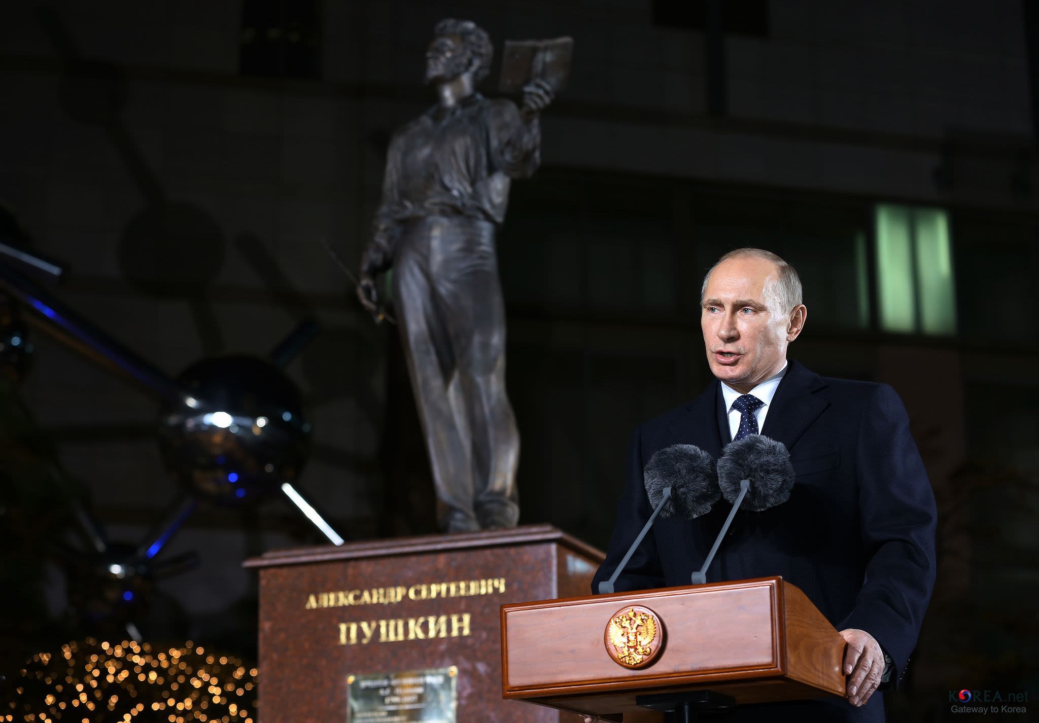 A middle-aged white man stands at a wooden lectern with two large, fluffy microphones in front of him, as he does a speech. There is a statue behind him with Russian writing on the base.