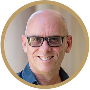 A headshot of a man wearing glasses smiling into the camera. You can only see part of his shoulders, he is wearing a blue shirt.