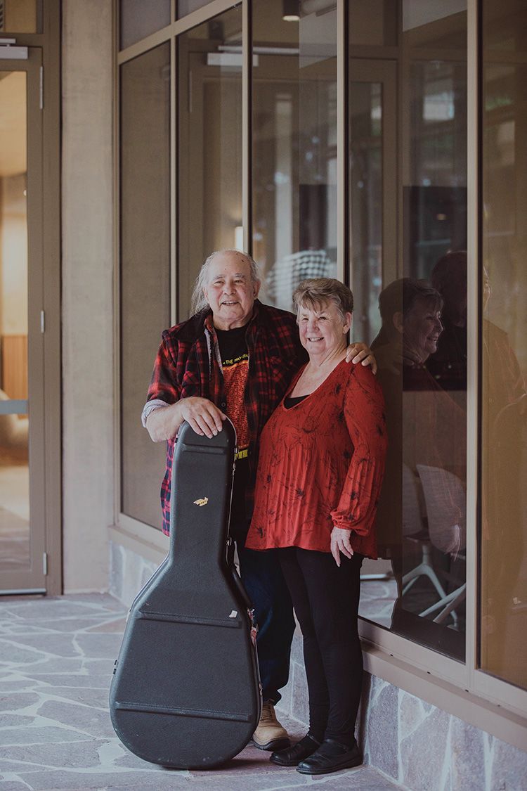 Kev Carmody stands with one hand on his guitar case, and the other around his wife's shoulders, and they break into laughter over the series of photos.