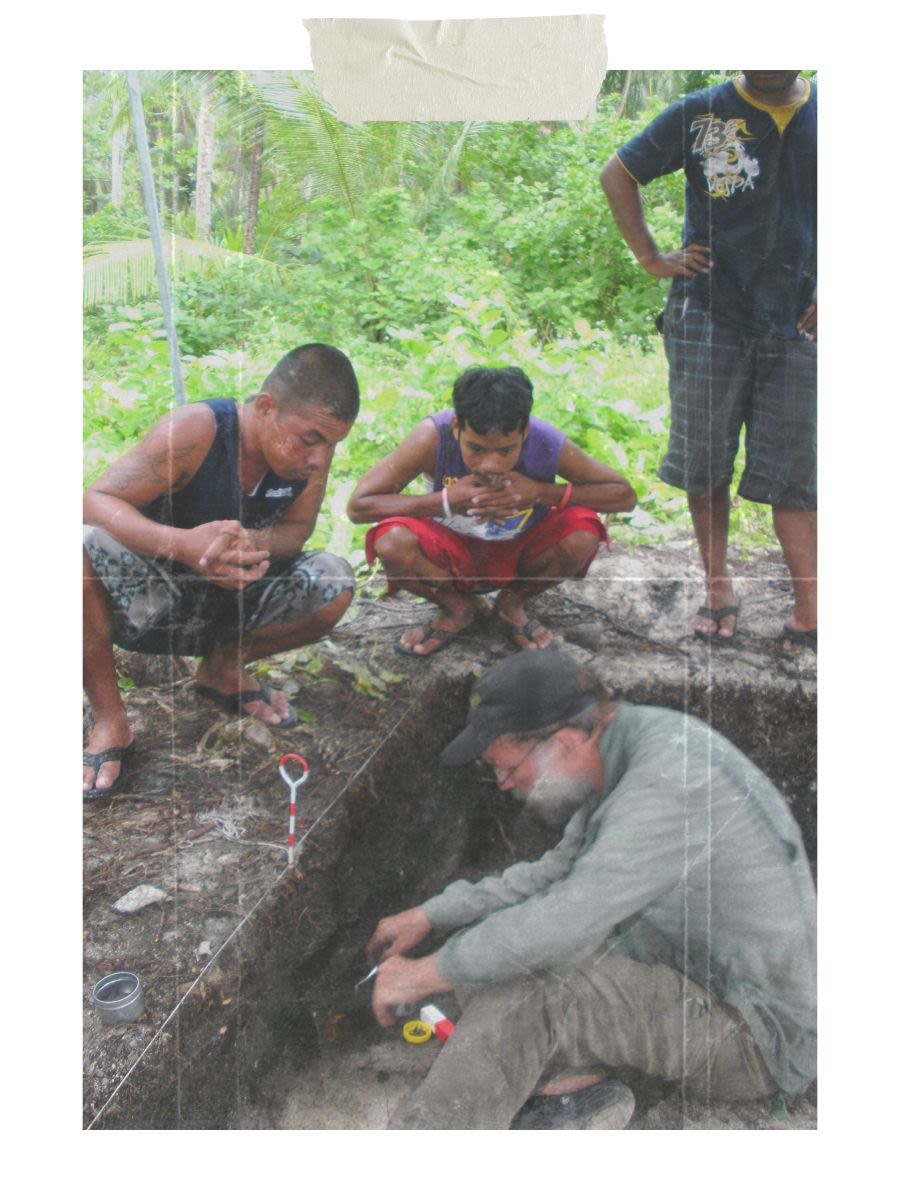 A man sits in a low trench examining something while three other men crouch and stand around him, looking at what he is doing. There is string around the rectangular dirt trench.
