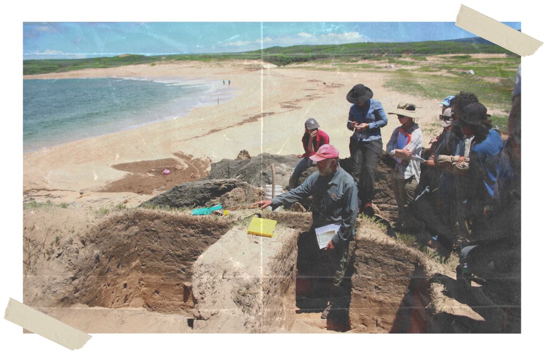 A man stands in a rectangular earth trench pointing at another trench. A group of students are gathered behind him, watching where he is pointing. The ocean and sandy beaches are behind them.