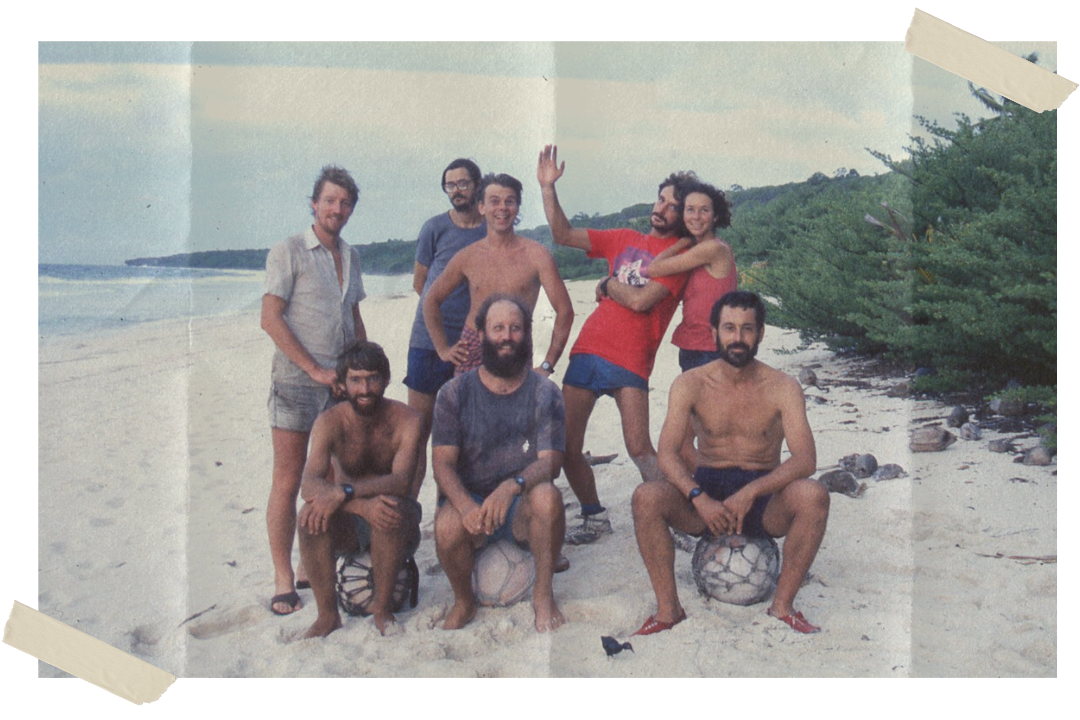 A group of six men and one woman on a sandy beach. The three in the front of the image are sitting on white balls covered in nets. They are smiling. The beach is empty and lined with shrubs.