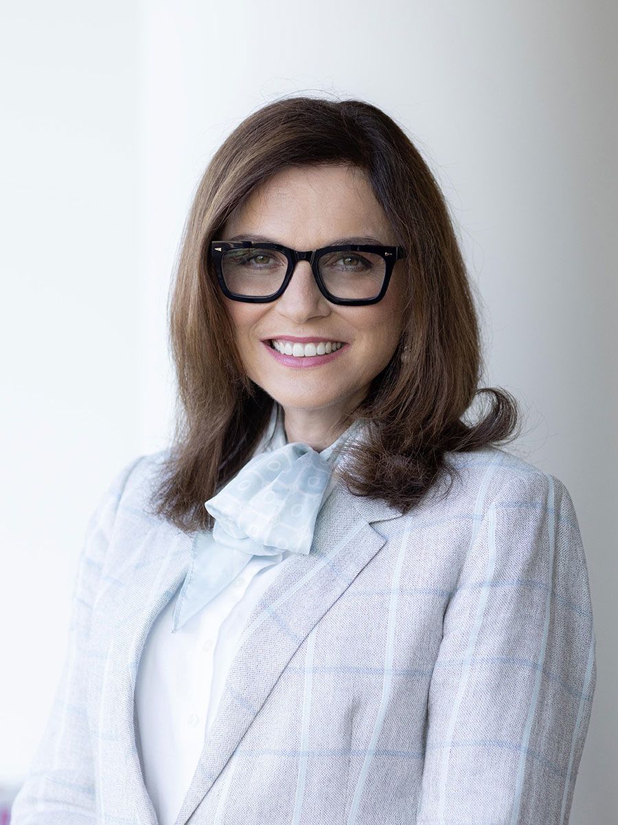 A smiling headshot of a woman with brown hair and glasses, wearing a blue check blazer.