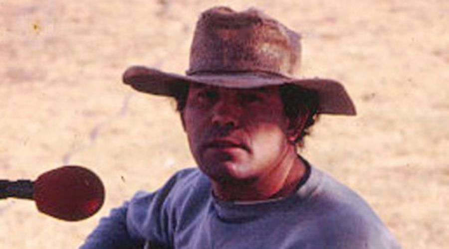 Photo of Kev Carmody as a young man. He has a hat on and there's a microphone next to him.
