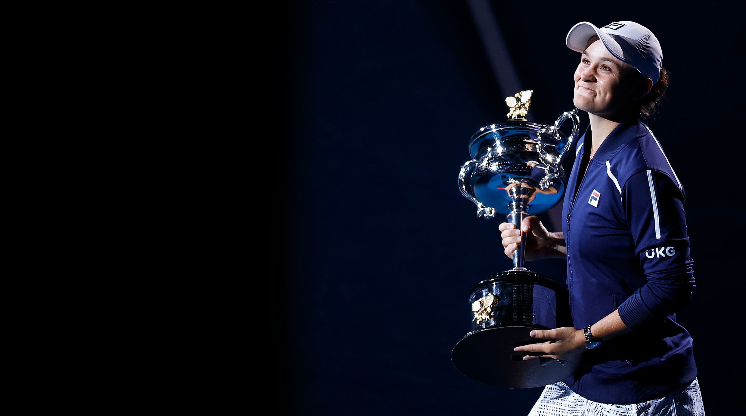 A woman holds a large silver trophy and smiles towards the camera. She is wearing a navy hoodie and a white cap.