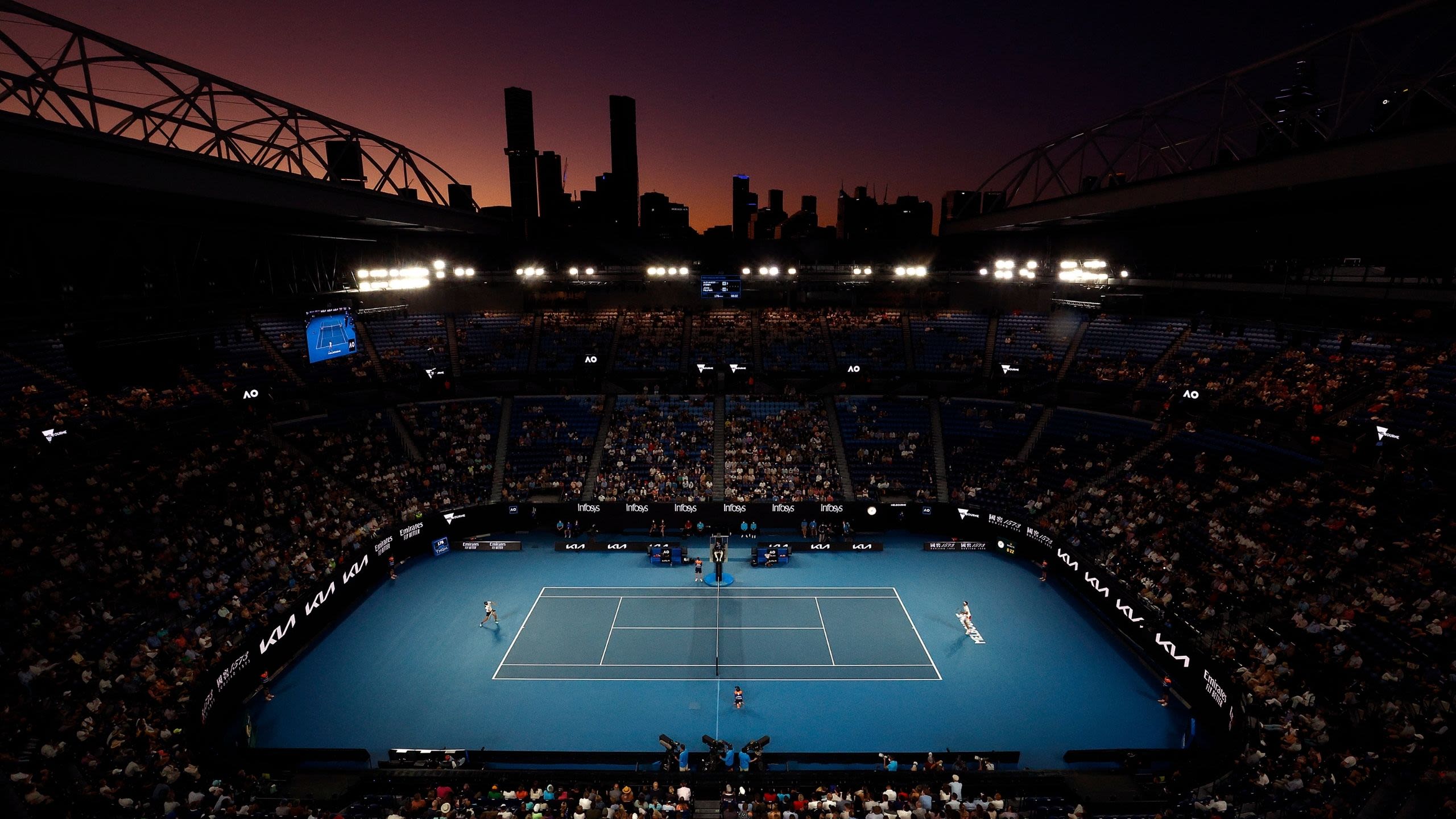 A tennis court is lit by two flood lights. Crowds fill the surrounding stadium, and in the background the sun is setting behind a silhouetted city skyline.