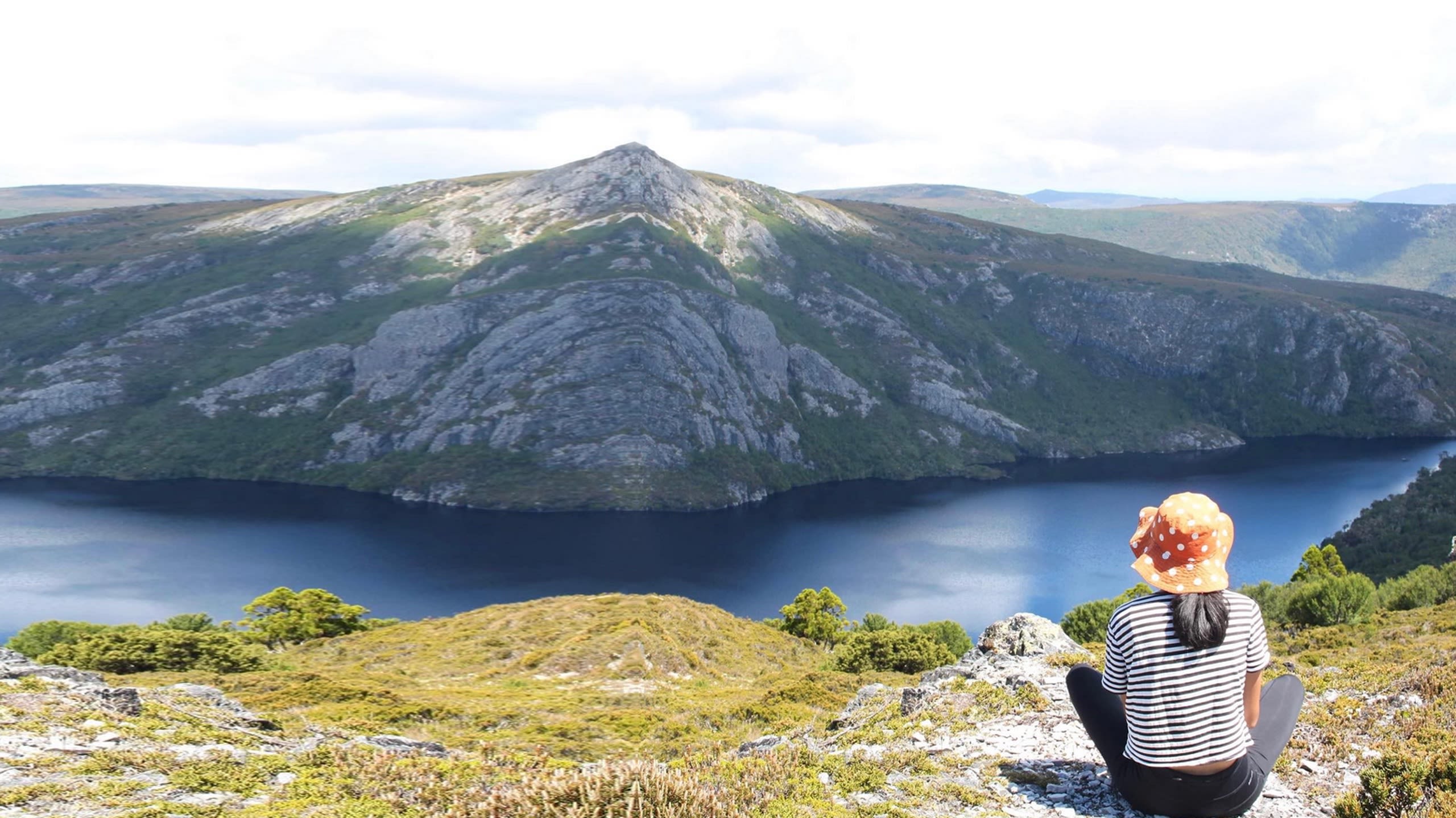 A woman in a colourful orange hat sits at a lookout overlooking a river and mountain range.