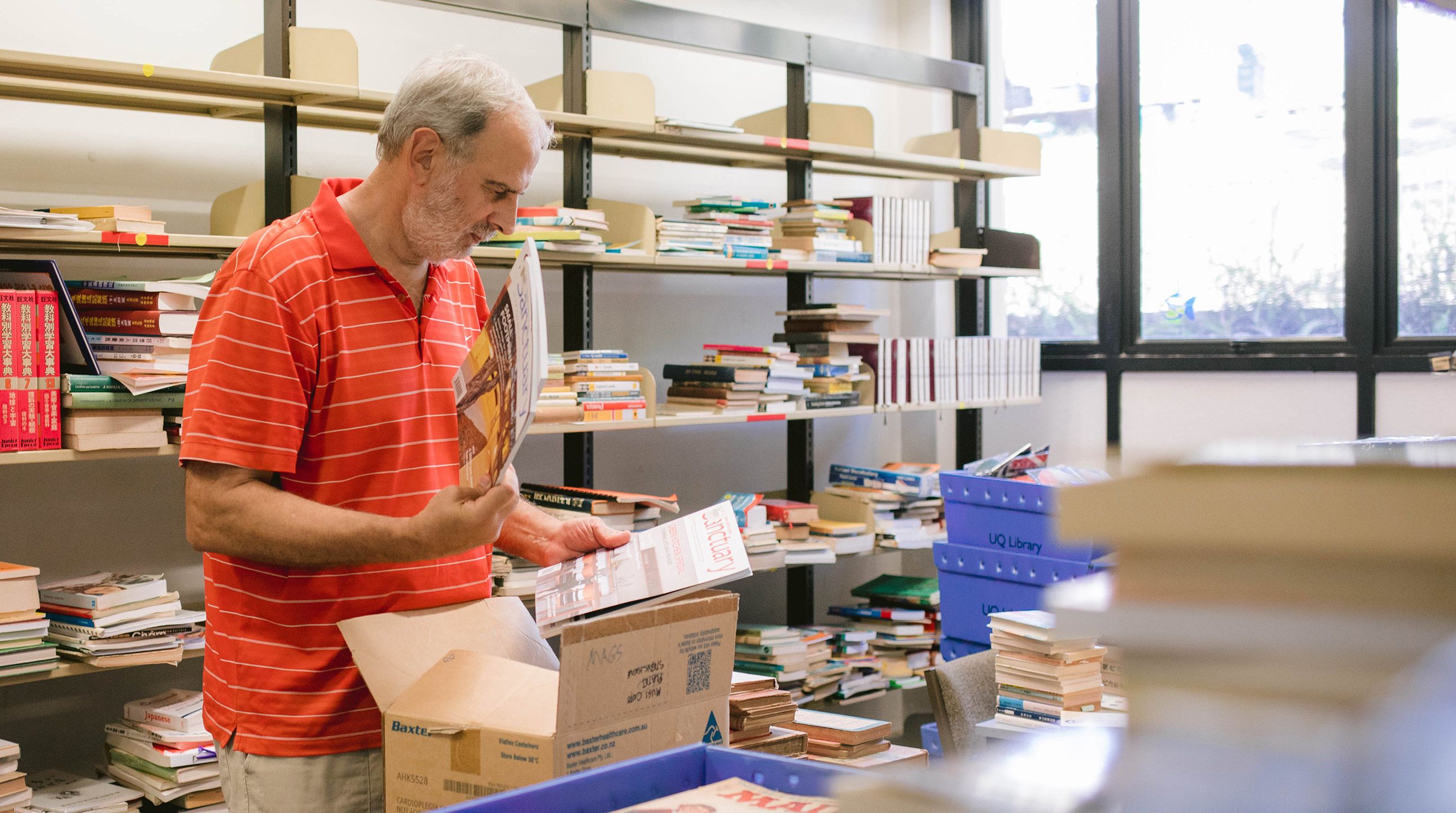A man in a red and white striped shirt with grey hair sorting through a box of books.