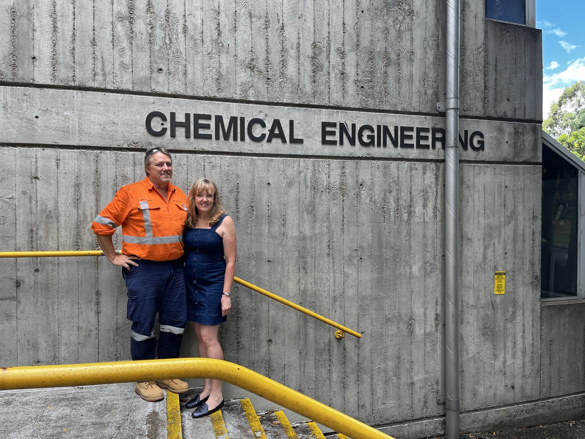 Wendy and Adrian pictured in front of the Chemical Engineering building at UQ St Lucia in February 2022. Adrian is dressed for work, wearing an orange high-vis shirt. Wendy is wearing a denim blue dress. They are both smiling at the camera.