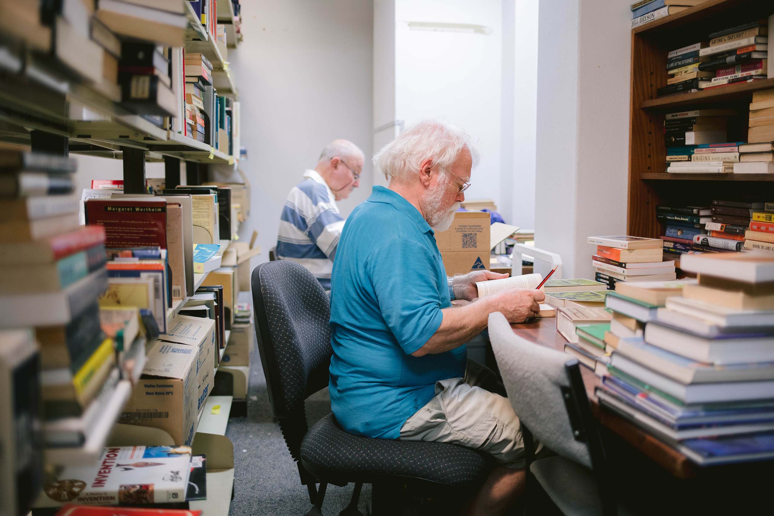 An elderly man sits at a desk surrounded by books, with another elderly man next to him. They are reading.