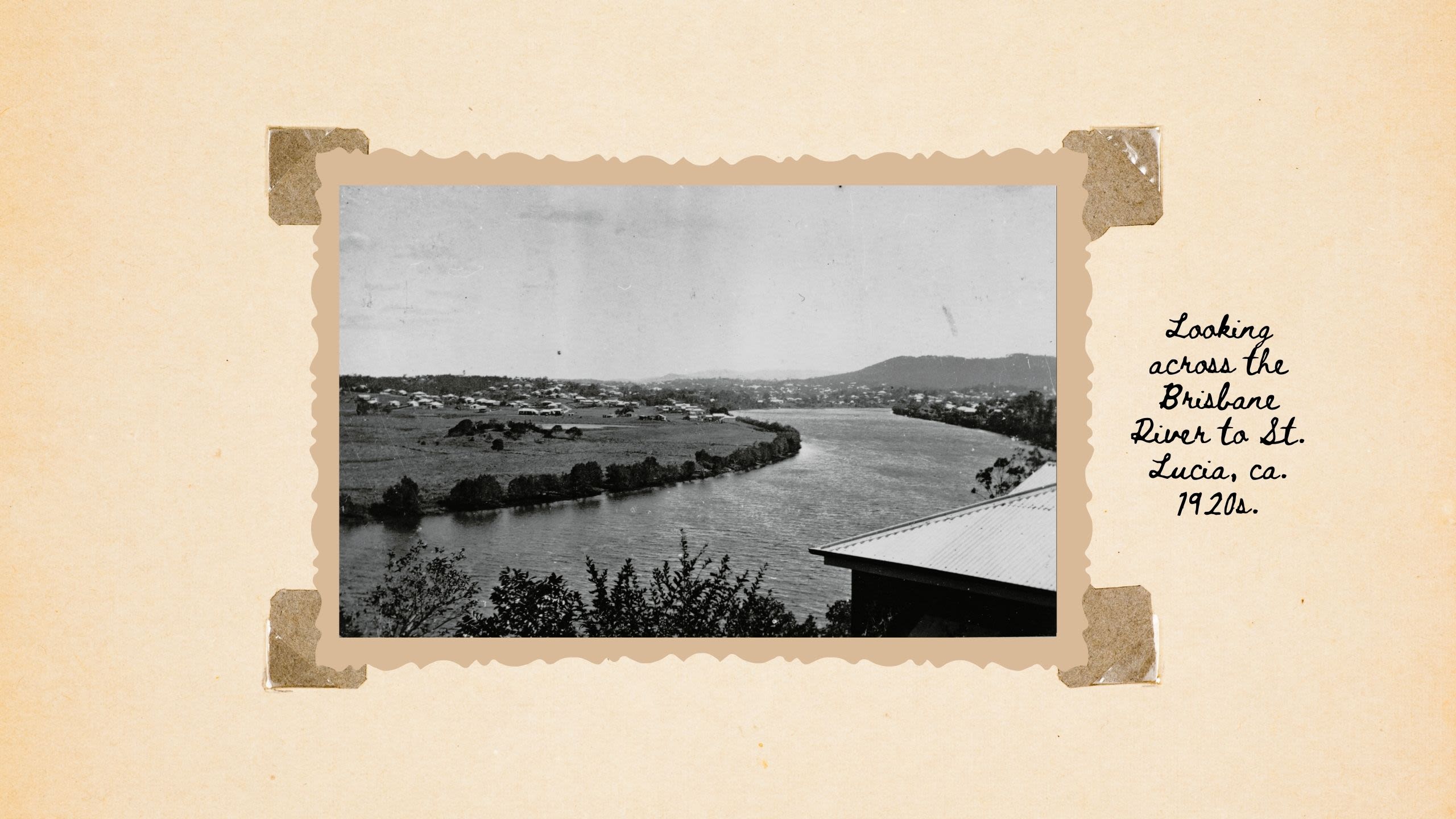 A black-and-white photo of a wide river, looking over the roof of a house. Green fields with few trees lie along the river's banks. The text reads 'Looking across the brisbane river to St Lucia ca. 1920s'
