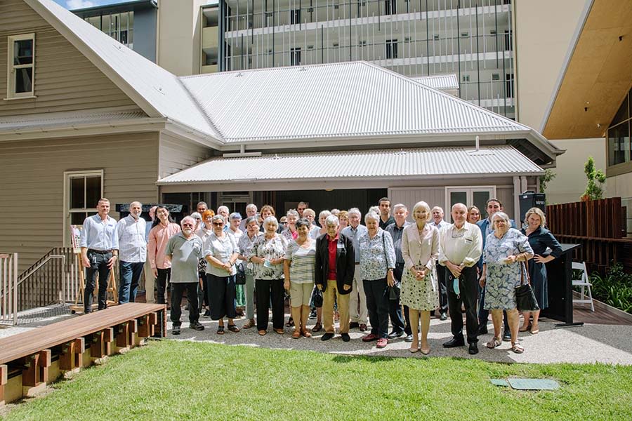 A group of around 25 people stand in front of a 'Queenslander' house. A high rise building is behind the house. There is a small green lawn in front of them and everybody is smiling.