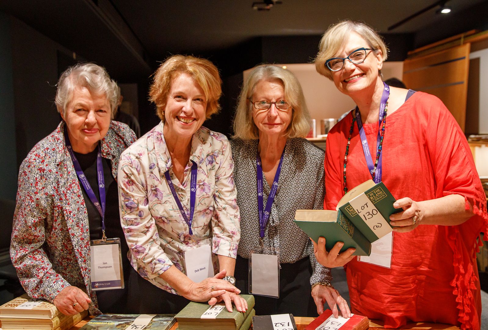 Four older women smile and stand next to each other wearing lanyards for the Book Fair. The women on the right is holding a book with a paper notice on it saying 'Lot 130'