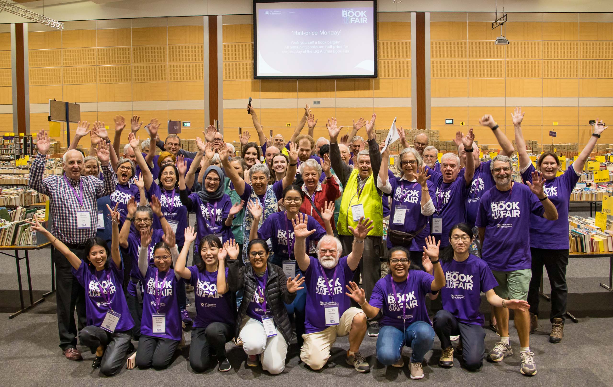 A group of 20 or more volunteers of varying ages - some elderly, some young students - wearing matching UQ Book Fair shirts. They are smiling and cheering as they put their hands up in celebration.