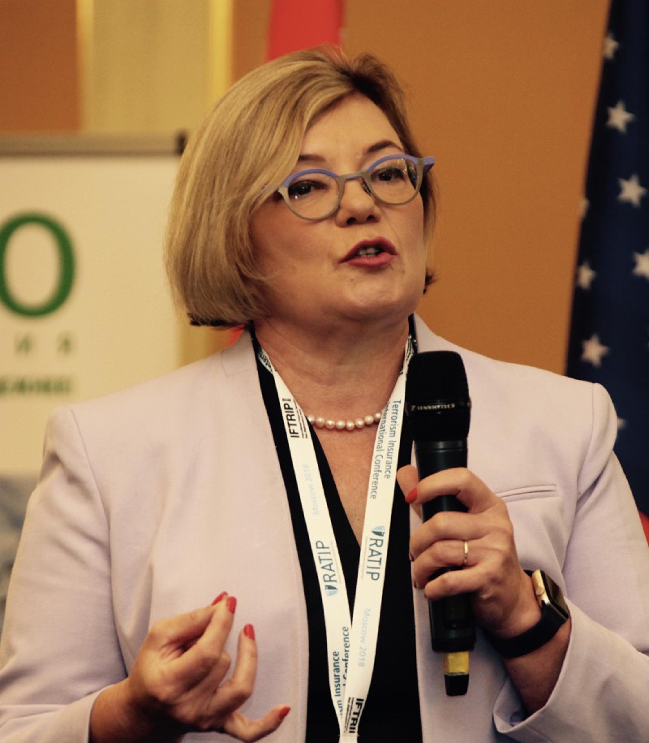 A woman with short blond hair and blue glasses speaks into a microphone..