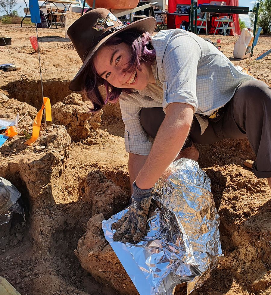 Nellie Pease at a dig site pressing down on the edge of a piece of silver foil that is covering something. She is wearing a broad-brimmed hat and gloves.