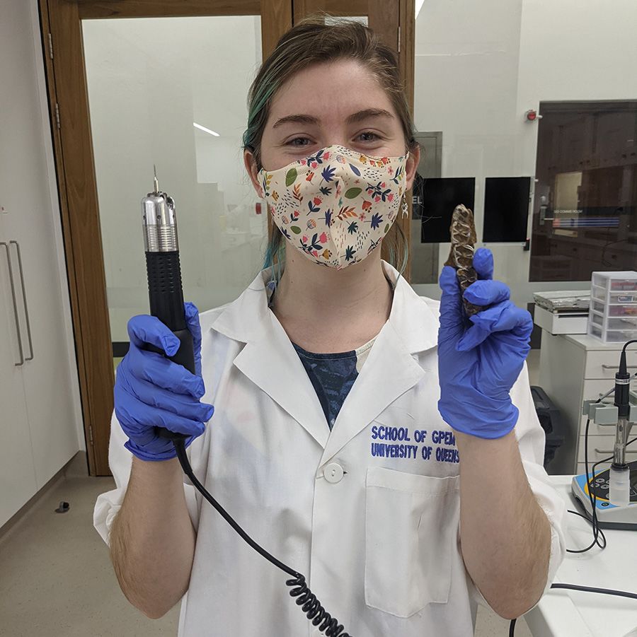 Nellie Pease wearing a cloth face mask and a white lab coat. She is holding a drill in one hand and a set of fossilised teeth in the other.