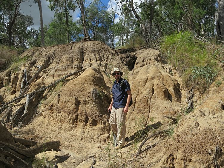 Dr Gilbert Price wearing weather-protective clothing standing in the sun at a fossil dig site. An embankment of sand and dirt is behind him.
