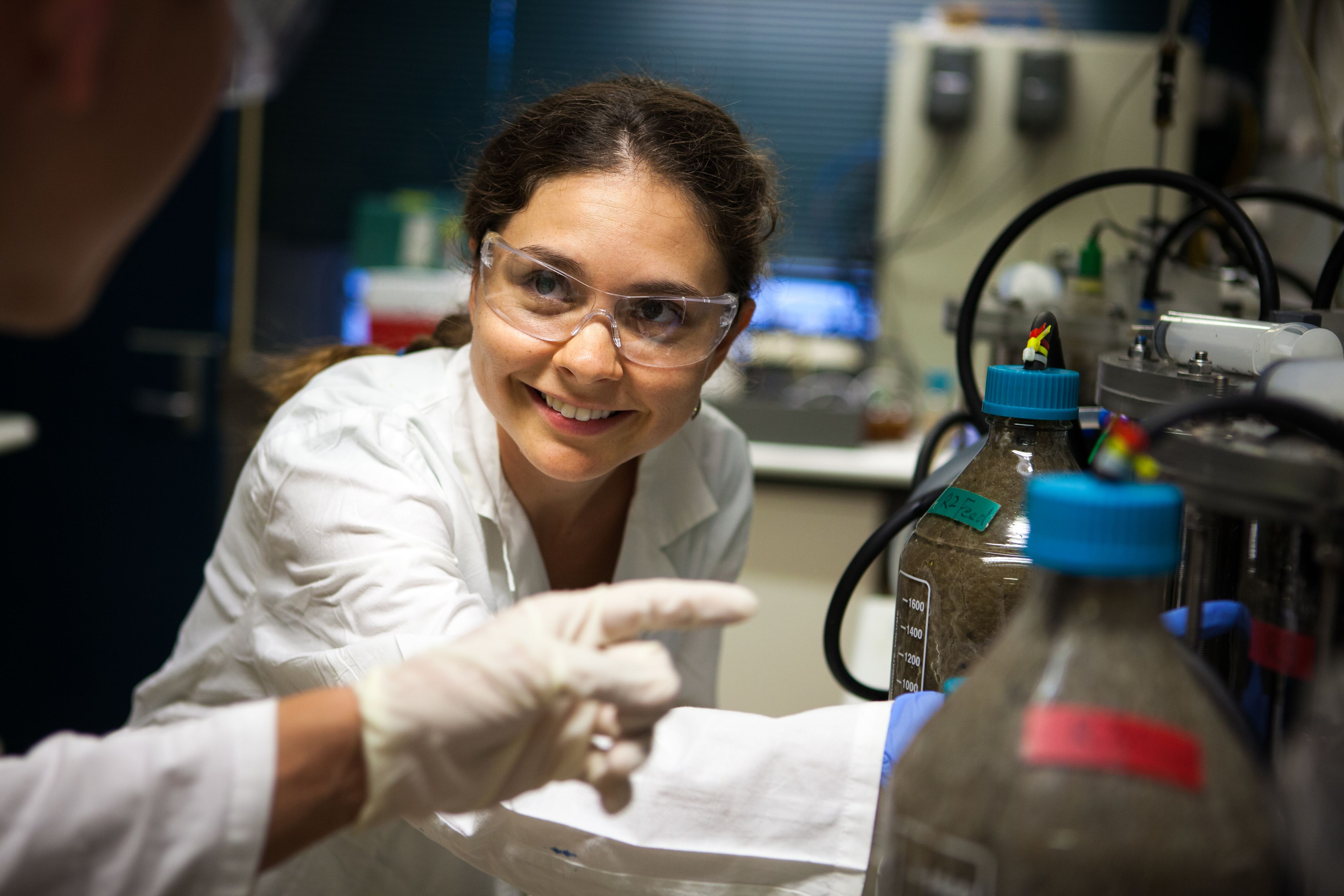A female scientist in a lab coat and safety glasses works alongside a colleague in a laboratory