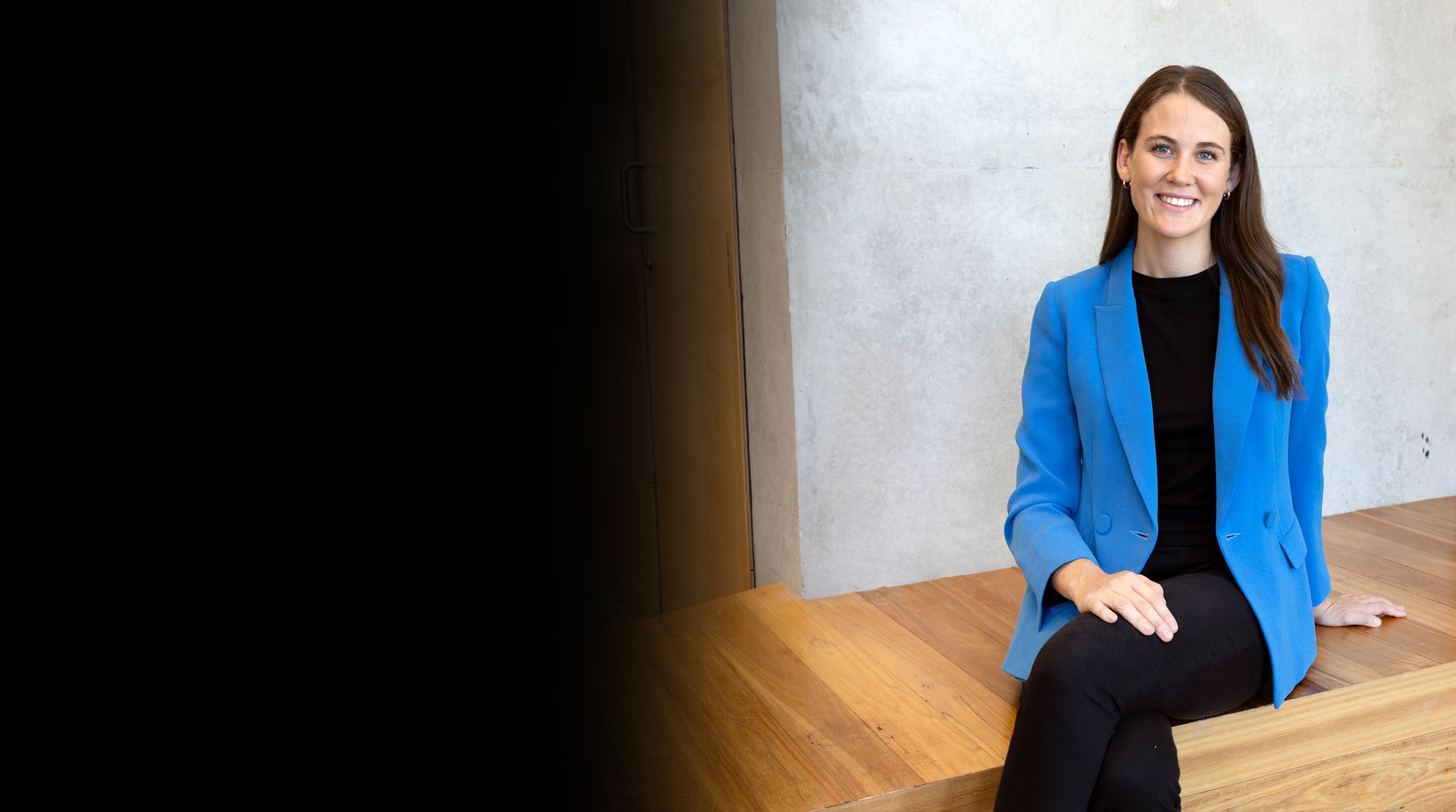 Hailey Brown sits on a wooden bench with her legs crossed. She is wearing black pants and singlet and a bright blue blazer and smiling at the camera.