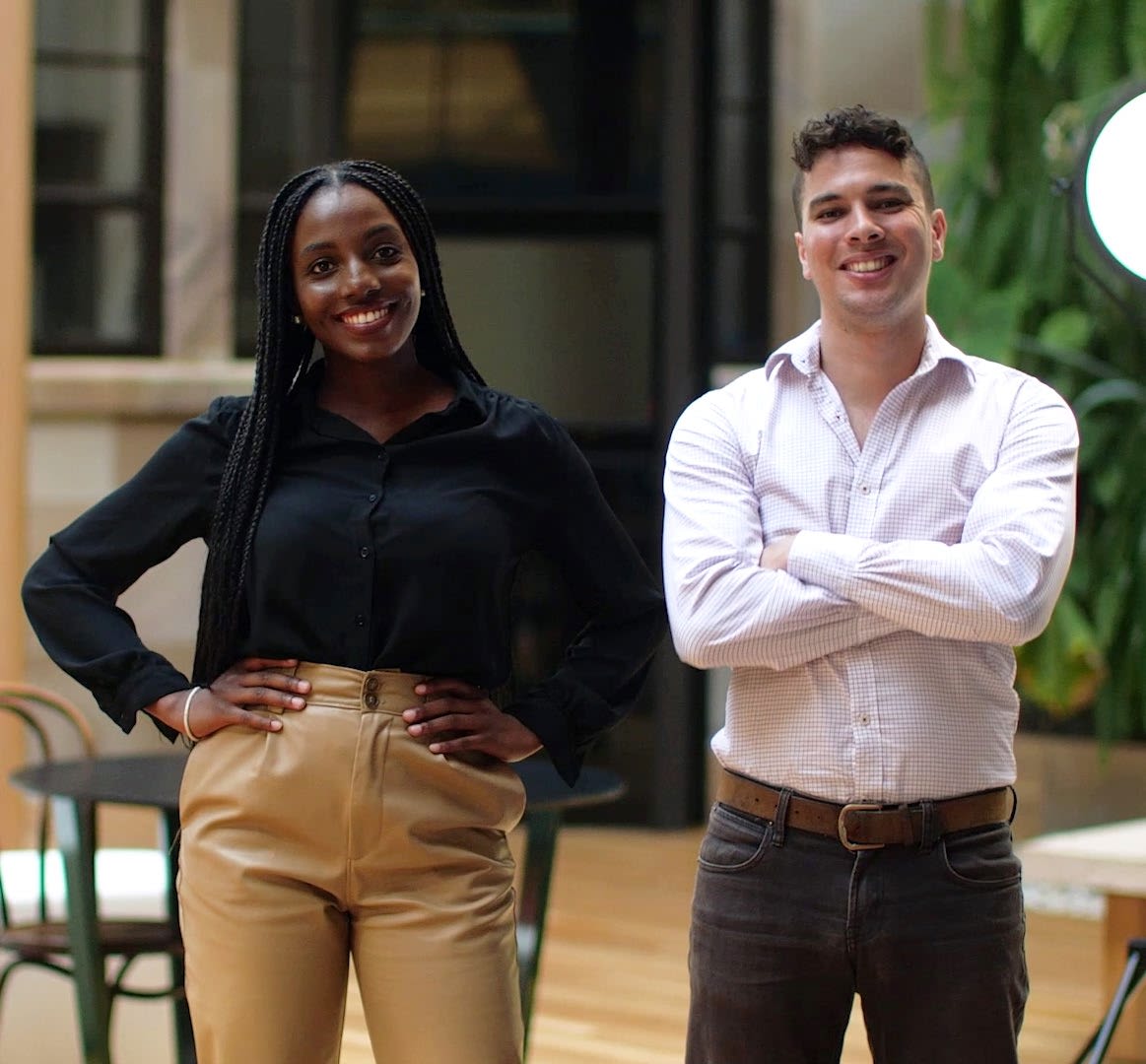Tadiwanshe Mhiti and Matthew Kairl stand in the UQ Change Institute. Tadiwanshe is wearing a black top with her hands on her hips and Matthew is wearing a white shirt with his arms folded.