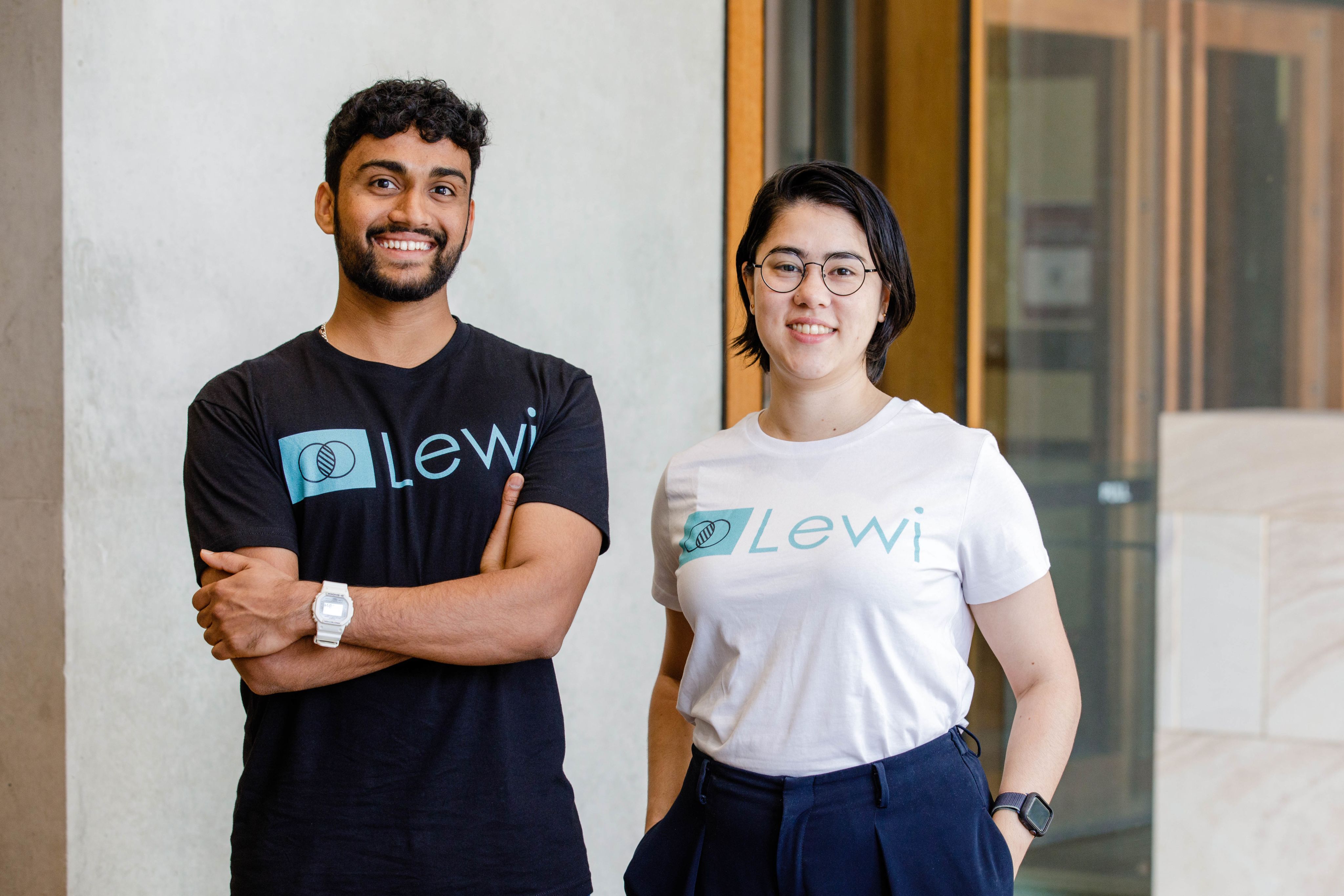 Lewi founders, Vishnu Avudainayagam and Geraldine Terada-Bellis stand for a photo. Vishnu is wearing a black t-shirt and Geraldine is wearing a white t-shirt - both have the company logo across the front.