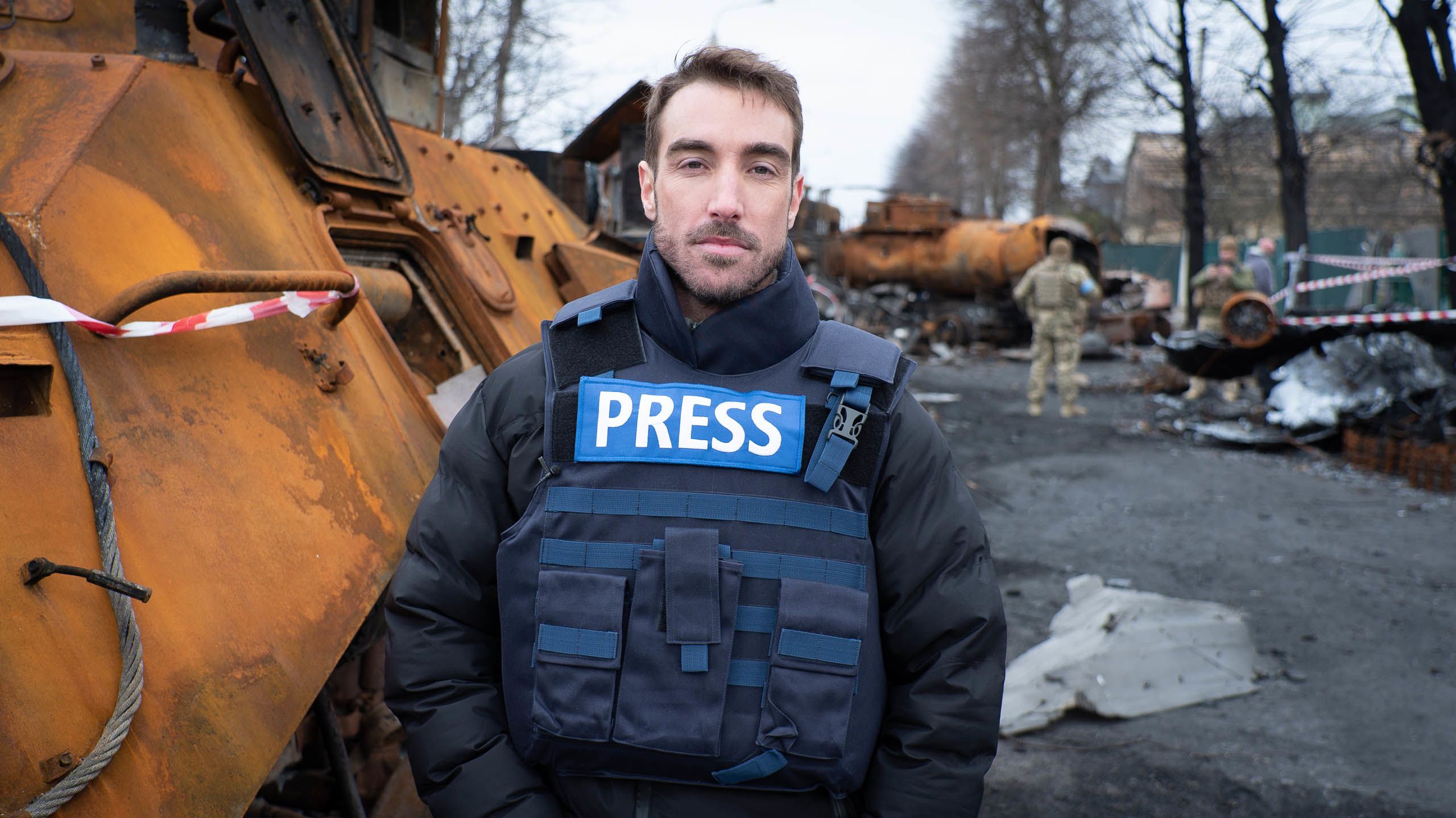A man in a blue press flak jacket standings in front of a road full of rubble.