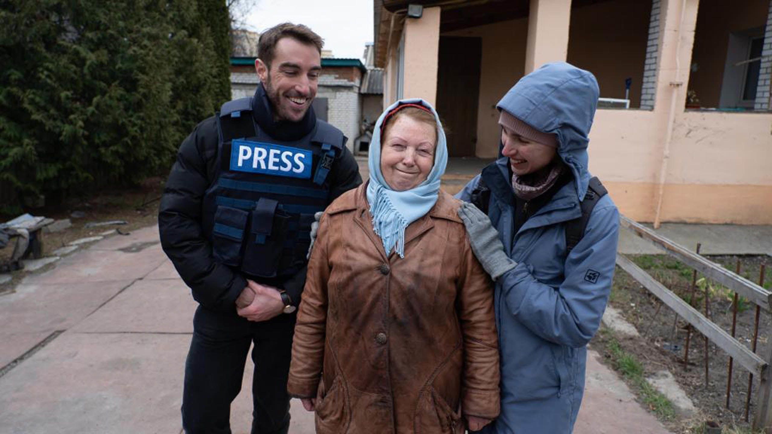 A man in a blue press flak jacket stands with two smiling women wearing coats.