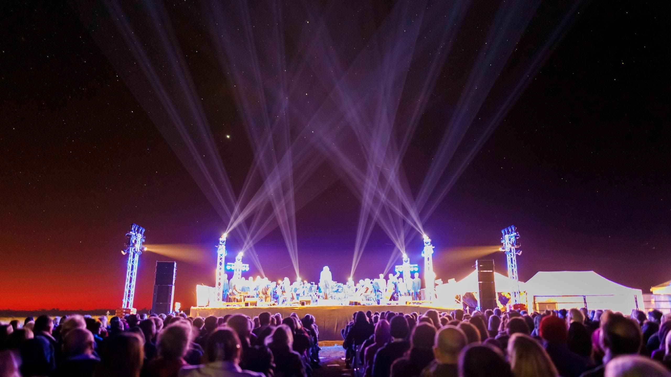 A large stage with orchestra performers lit up under the starry night sky, with the audience in front of them