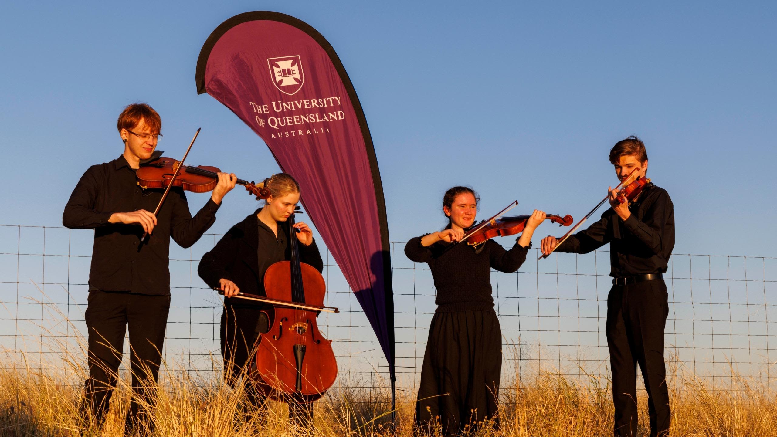 Four musicians in black clothes stand in a field holding instruments