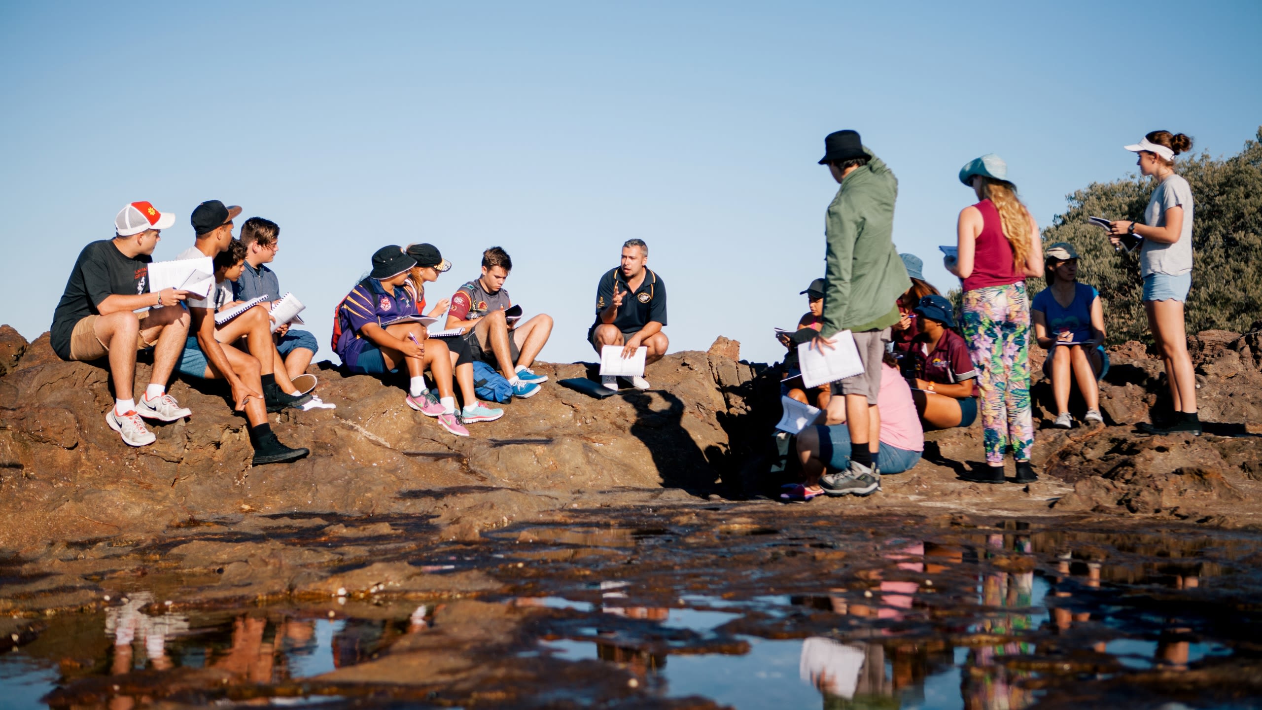 A large group of young people sit on rocks outside with a teacher