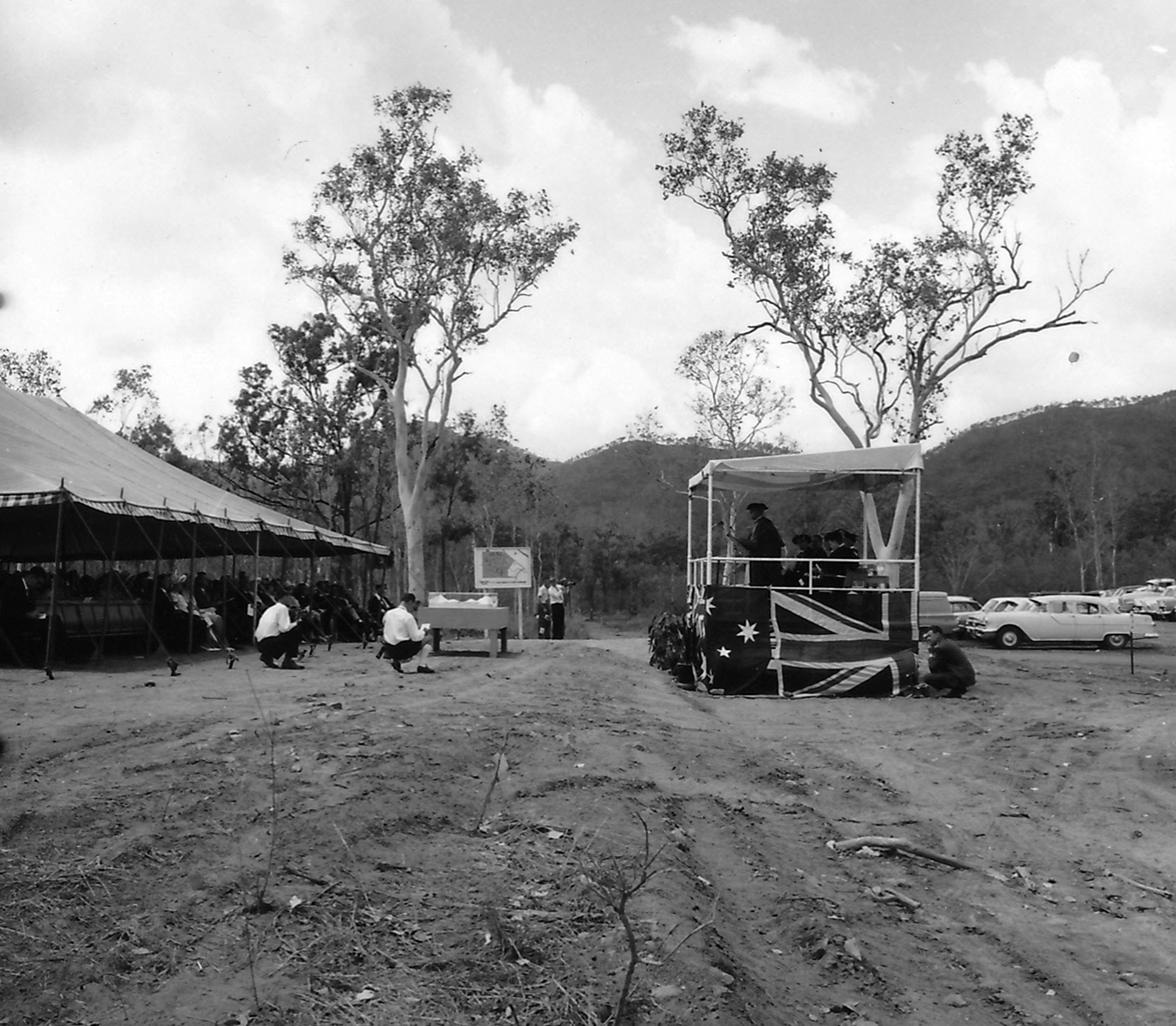 A black and white photo of a marquee and a small podium opposite one another. The podium has the Australian flag wrapped around it. There is a speaker at the lecturn on the podium addressing the audience in the marquee. 