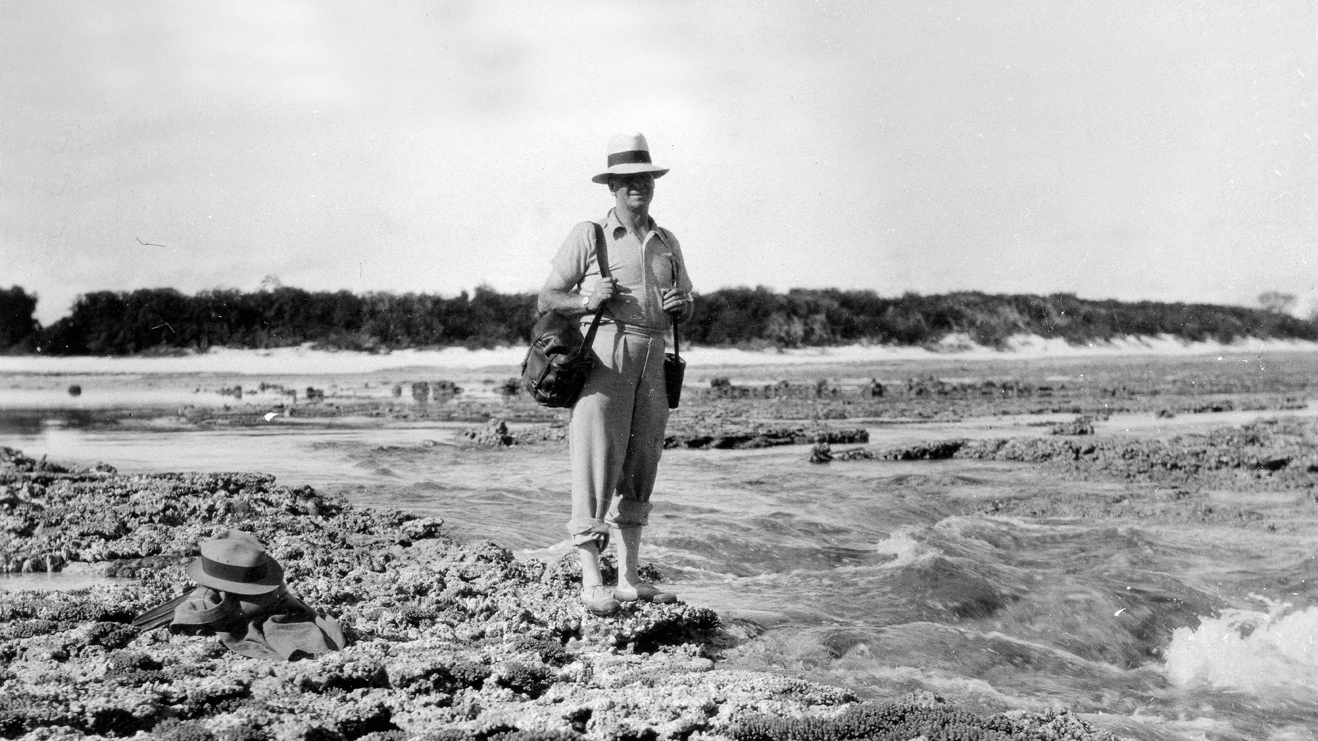 A black and white photo of a man with a straw sun hat and carrying two over-the-shoulder satchels with his pants rolled up around his ankles, wearing reef shoes standing on the reef rocks covered in coral